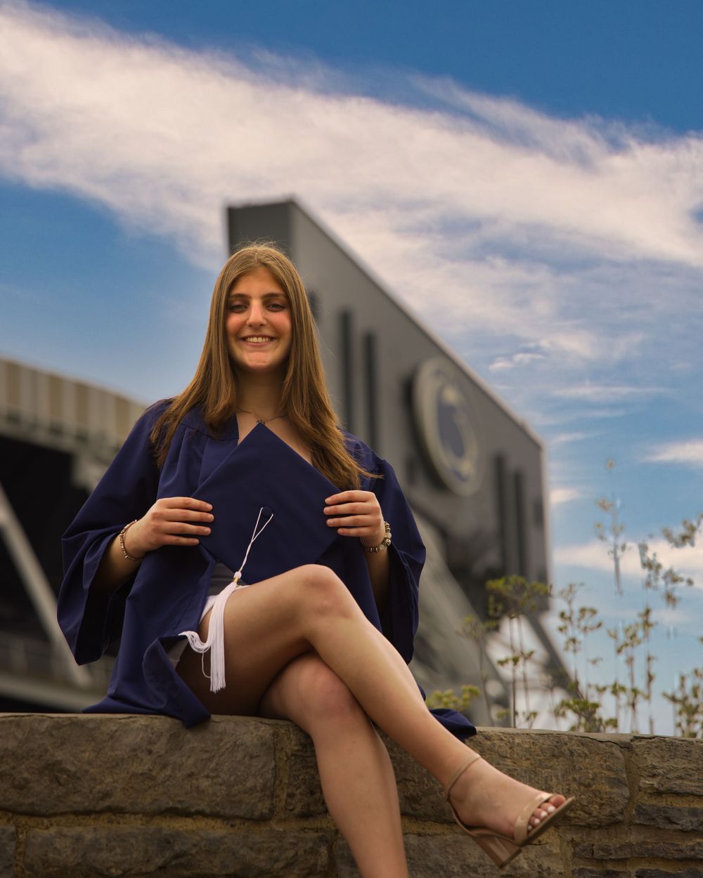 Penn State graduate in cap and gown sitting on the stone wall below Beaver Stadium
